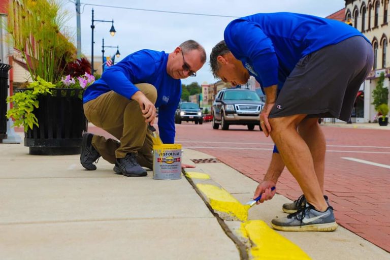 Two Homestead Saving Bank workers bending down to repaint the border of the sidewalk yellow for Community Unity Day.
